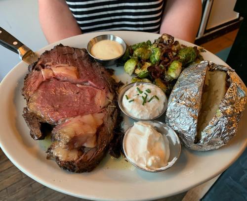 A plate featuring a large slice of prime rib, baked potato, Brussels sprouts, and dipping sauces.