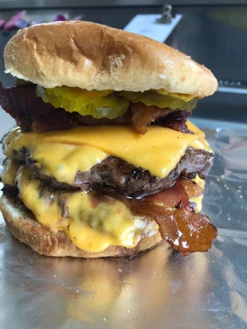 A towering cheeseburger with three patties, melted cheese, bacon, pickles, and a soft bun, resting on foil.