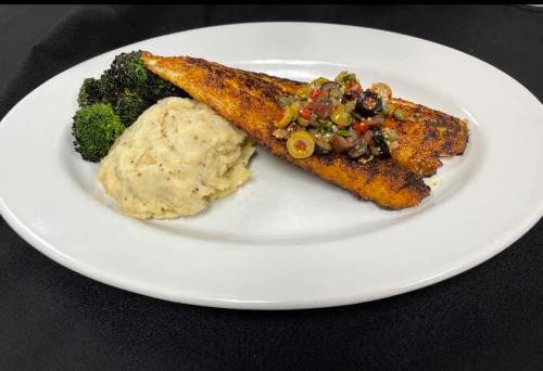 A plate featuring seasoned fish fillet topped with olive salsa, served with mashed potatoes and steamed broccoli.