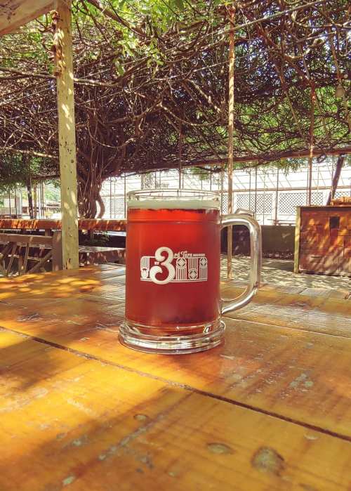 A glass mug of dark beer sits on a wooden table under a shaded outdoor area.