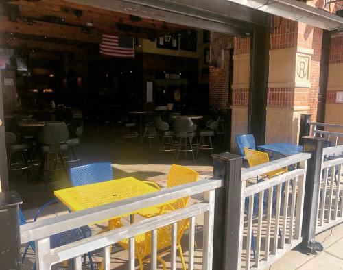 Outdoor seating with yellow and blue tables in front of a restaurant, partially open with a view of empty chairs inside.