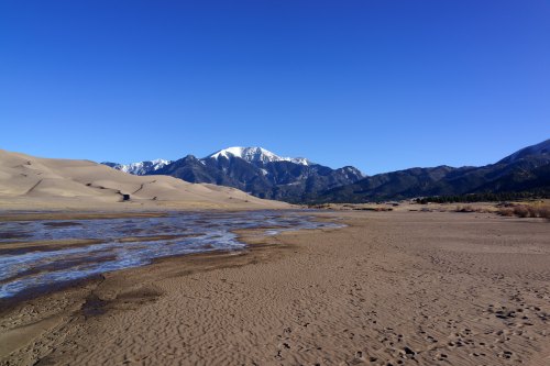 A serene landscape featuring sandy dunes, a winding stream, and snow-capped mountains under a clear blue sky.