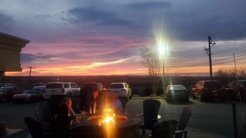 A group of people sits around a fire pit at sunset, with a colorful sky and parked cars in the background.