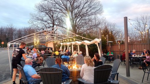 Outdoor dining scene with people enjoying a meal around a fire pit under a tent, surrounded by trees and evening sky.