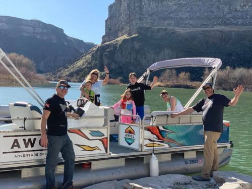 A group of six people wave from a colorful boat on a river, surrounded by rocky cliffs and greenery.
