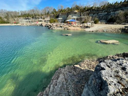 A serene lake with clear green water, rocky shores, and a small building in the background surrounded by trees.