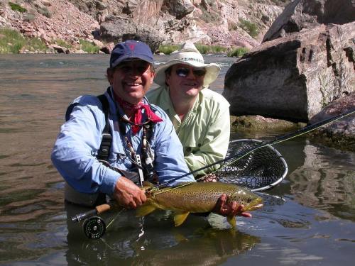 Two men stand in a river, one holding a large trout, with fishing gear and rocky terrain in the background.