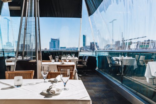 A modern restaurant with outdoor seating, featuring tables set for dining and a view of the city skyline.