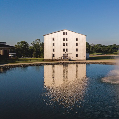 A white barn reflected in calm water, surrounded by greenery and a clear blue sky.
