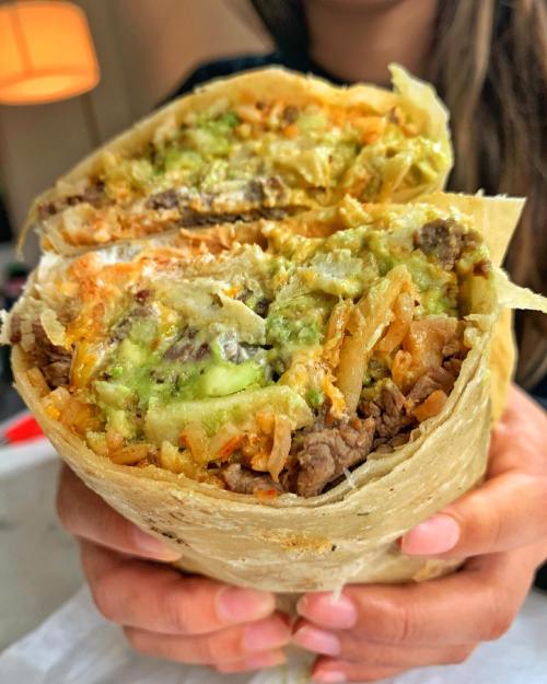 A close-up of a large burrito filled with beef, rice, and guacamole, held by a person.