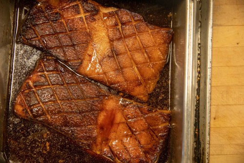 Two pieces of glazed, roasted meat with a crisscross pattern, resting in a dark, flavorful sauce.