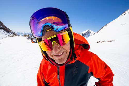 A smiling skier in a bright orange jacket and colorful goggles stands on a snowy mountain slope under a clear blue sky.