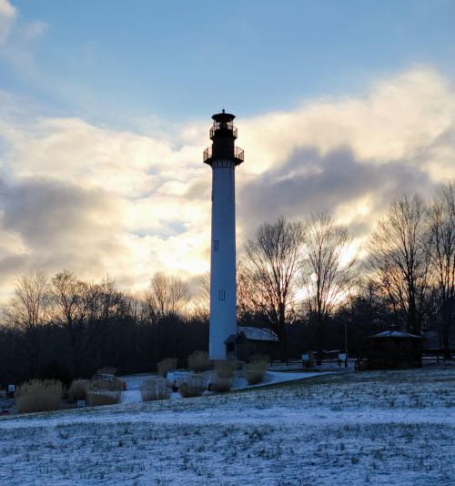 A tall lighthouse stands against a cloudy sky, surrounded by snow-covered ground and bare trees.