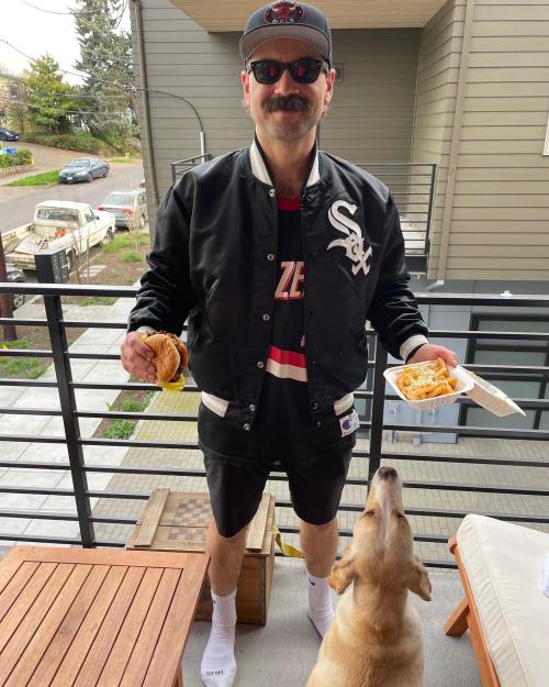 A man in a black jacket and sunglasses holds a sandwich and a takeout box, with a dog nearby on a balcony.