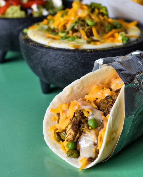 A close-up of a taco filled with meat, cheese, and vegetables, with a bowl of nachos in the background.