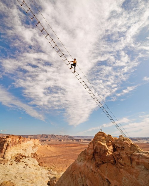 A person climbs a tall, narrow ladder suspended over a vast desert landscape under a blue sky with clouds.