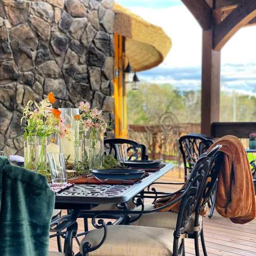 A beautifully set outdoor dining table with flowers, glassware, and a cozy blanket, against a stone wall backdrop.