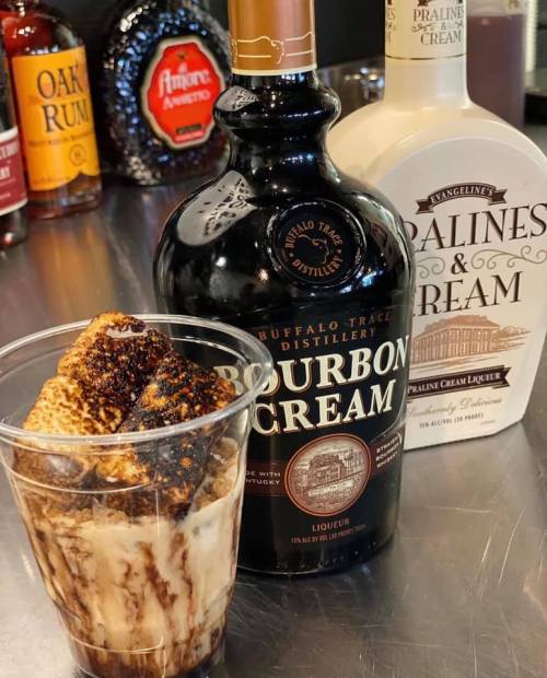 A cup with a creamy dessert topped with chocolate sauce, alongside bottles of Bourbon Cream and Praline Cream liqueurs.
