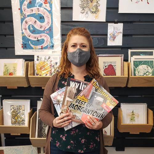A woman wearing a mask holds magazines in a shop with framed art and prints on the wall behind her.
