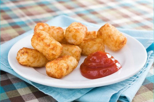 A plate of golden-brown tater tots served with a side of ketchup on a blue napkin, set on a checkered tablecloth.