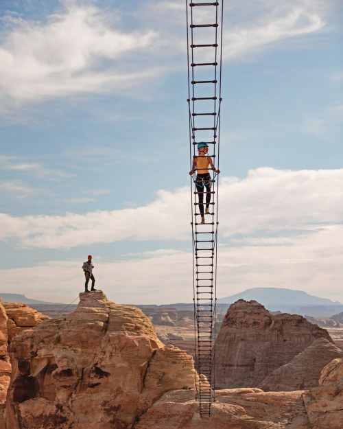 A person climbs a tall metal ladder on a rock formation, while another stands on a nearby cliff, against a scenic backdrop.