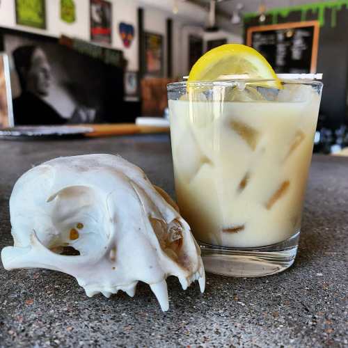 A glass of creamy drink with ice and a lemon slice next to a small animal skull on a textured surface.