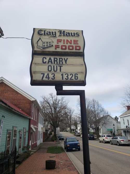 Sign for Clay Haus Fine Food displaying "Carry Out" and a phone number, with a street view of nearby buildings.