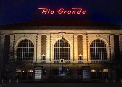 Historic building with large windows, illuminated at night, featuring a "Rio Grande" sign and an American flag.