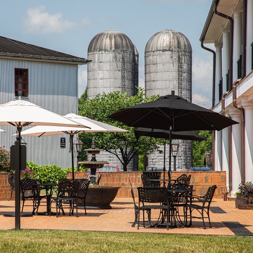 A patio with black and white umbrellas, tables, and chairs, overlooking two silos and greenery in the background.