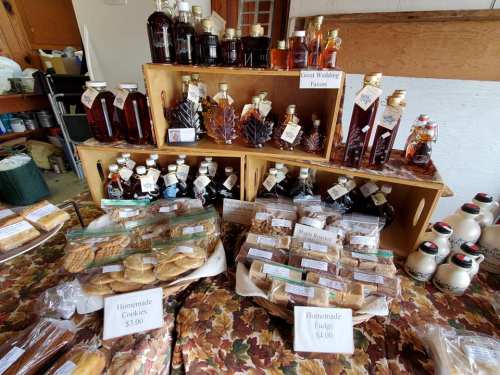 A display of homemade goods featuring bottles of syrup, fudge, and cookies, with prices labeled on each item.