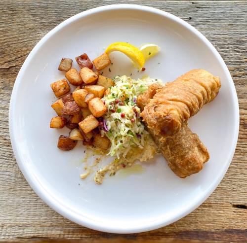 A plate with fried fish, coleslaw, roasted potatoes, and lemon wedges on a wooden table.
