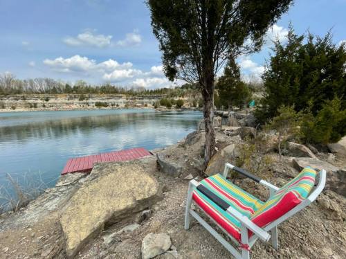 A colorful chair by a calm lake, surrounded by rocks and trees, with a wooden dock in the background under a cloudy sky.