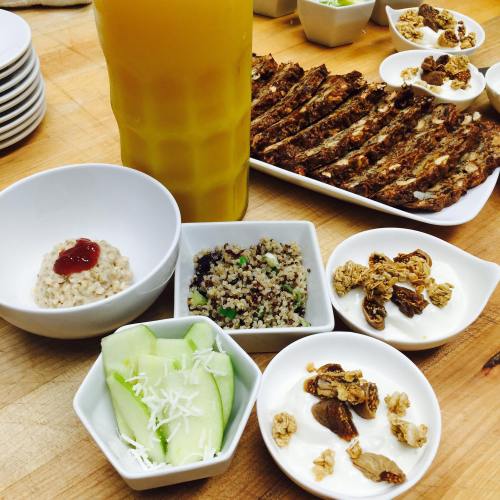A spread of food including ribs, quinoa salad, cucumber slices, granola, and a drink on a wooden table.