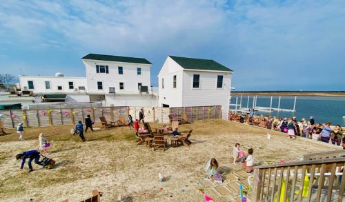 Children search for Easter eggs in a grassy area near waterfront houses, with colorful decorations and a festive atmosphere.