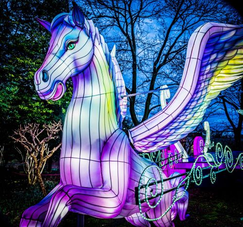 A colorful, illuminated winged horse sculpture against a twilight sky, surrounded by bare trees.