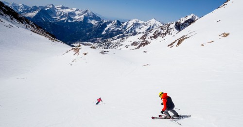 Two skiers navigate a snowy slope with mountains in the background under a clear blue sky.