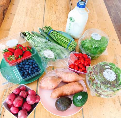 A variety of fresh produce including strawberries, blueberries, tomatoes, sweet potatoes, avocados, and greens on a wooden table.