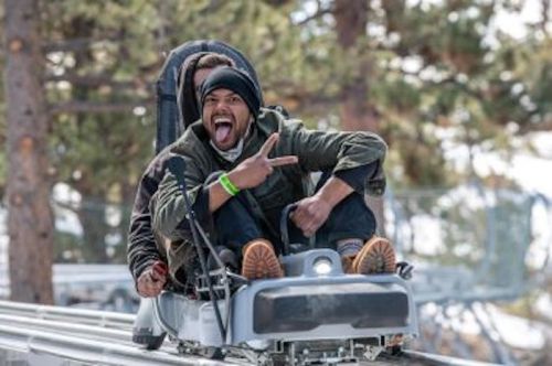 Two people on a roller coaster, one making a peace sign and sticking out their tongue, surrounded by trees.