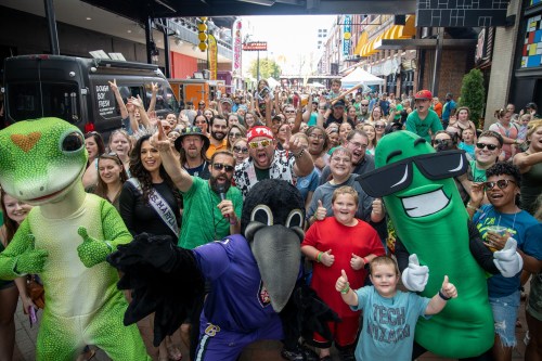 A lively crowd celebrates with mascots, smiling and giving thumbs up at a festive outdoor event.