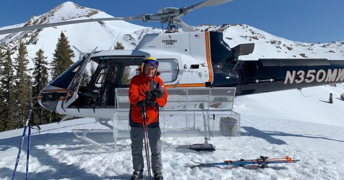 A skier in an orange jacket stands next to a helicopter on a snowy mountain landscape.
