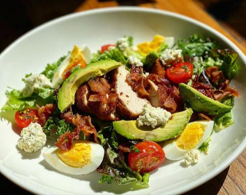 A vibrant salad with grilled chicken, bacon, avocado, cherry tomatoes, hard-boiled eggs, and crumbled cheese on greens.