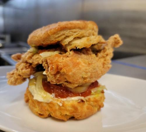 A stacked biscuit sandwich with fried chicken, pickles, and sauce on a white plate.