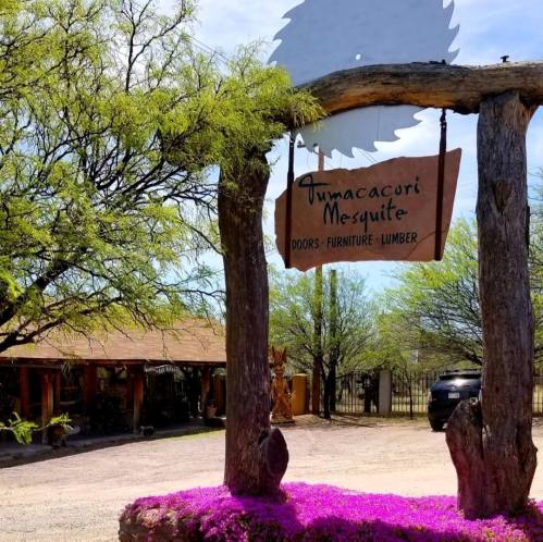 Sign for Tumacacori Mesquite, featuring doors, furniture, and lumber, surrounded by vibrant purple flowers and greenery.