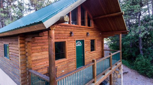 A rustic log cabin with a green roof, featuring a porch and surrounded by trees.