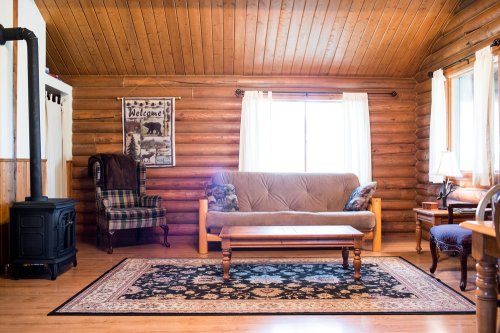 Cozy log cabin interior with a futon, wooden furniture, a patterned rug, and large windows letting in natural light.