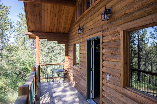 A wooden cabin porch with a table and chairs, surrounded by trees and natural scenery.
