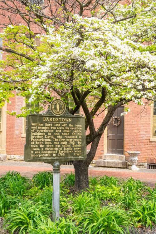 A historical marker in Bardstown, Kentucky, beside a flowering tree and brick building, detailing the town's founding.