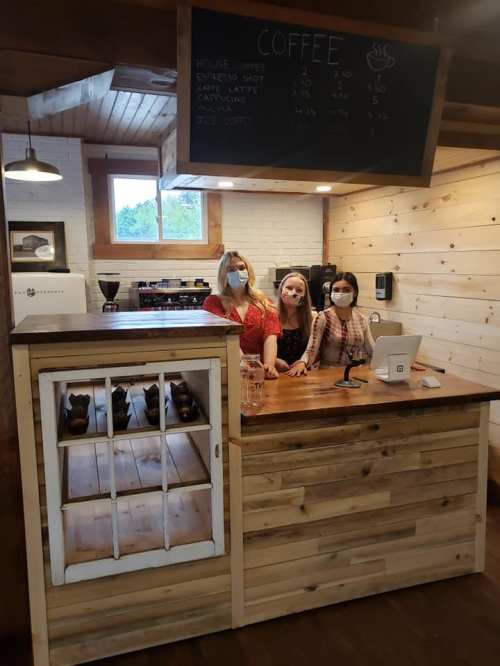 Three women wearing masks stand behind a wooden coffee counter in a cozy café setting. A menu is displayed above.