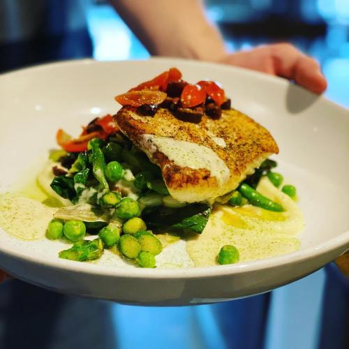 A plate of grilled fish topped with tomatoes, served on a bed of greens and peas, garnished with a creamy sauce.