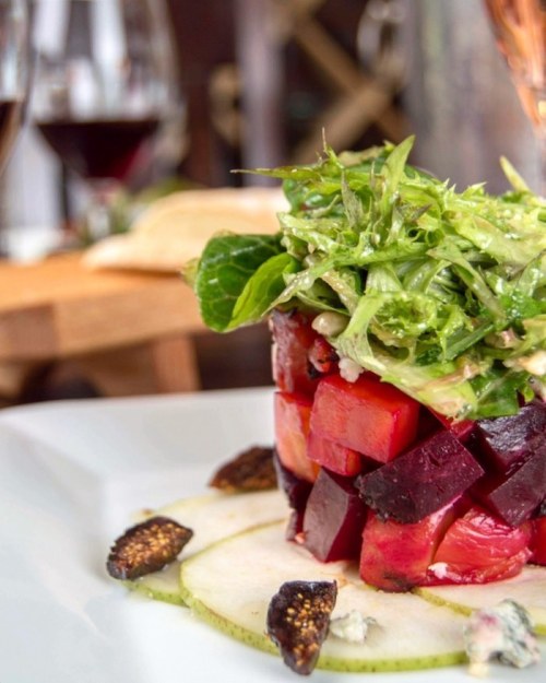 A colorful salad tower with beets, greens, and figs, served on a white plate with wine glasses in the background.